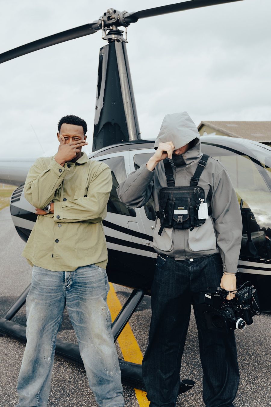 Oliver in a hooded jacket holding a cinema camera next to an artist in front of a helicopter