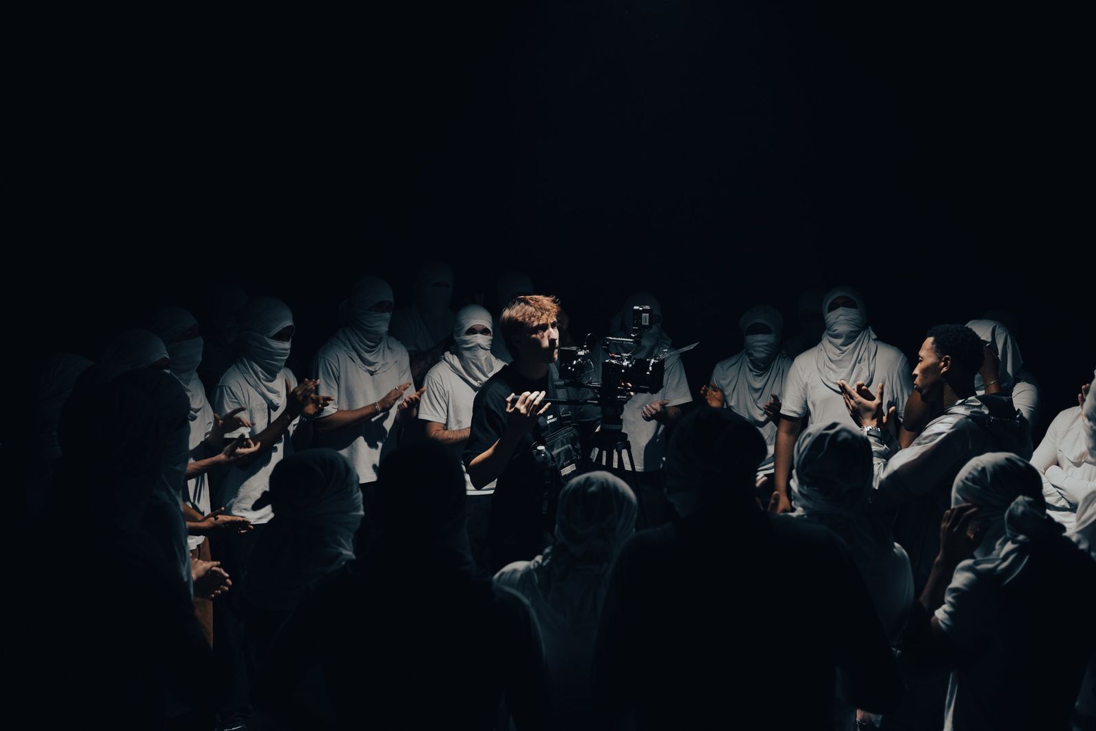 Oliver filming with a camera on a tripod, surrounded by a crowd in white face coverings on a dark set